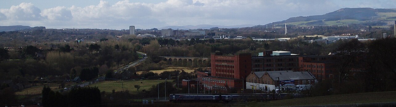 View of Glenrothes seen in its landscape setting from a nearby cemetery. A train is leavng nearby Markinch Station on the East Coast Mainline. Glenrothes town centre with the numerous taller residential and office buildings can be seen in the centre of the image. The River Leven Bridge provides a stark white vertical emphasis on the right side of the image. The Lomond Hills regional park and rolling countryside form the backdrop on the horizon.